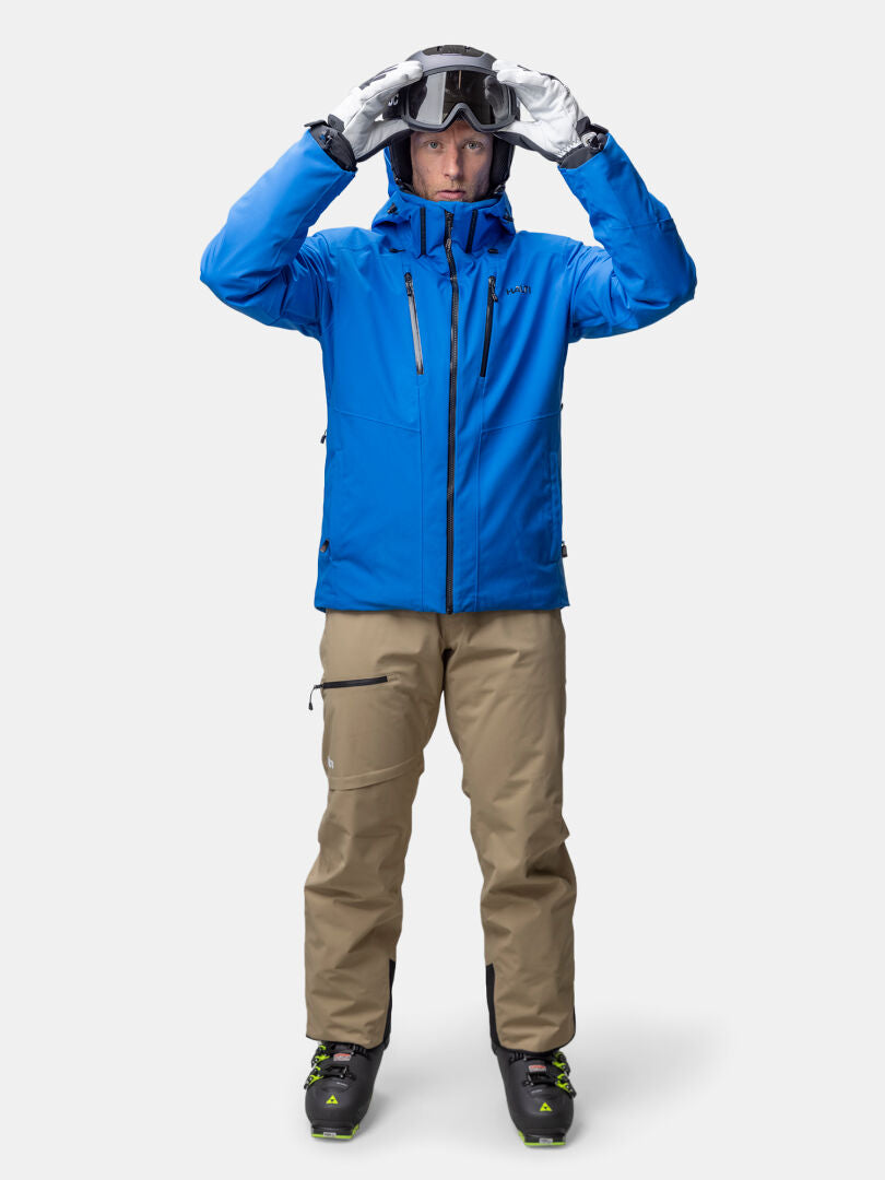 Eine Person in einer blauen Halti Vertica DX Skijacke Herren und einer hellbraunen Hose passt die Skibrille vor einem schlichten Hintergrund an.