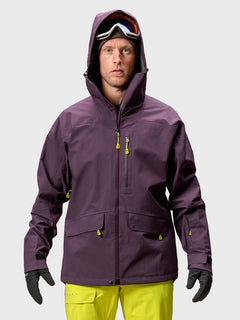 Ein Mann trägt eine Halti Settler 3L DX Ski Jacke Herren in lila, gelbe Hosen, Handschuhe und eine Skibrille vor einem schlichten Hintergrund.