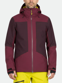 Ein Mann trägt eine kastanienbraune Halti Planker DrymaxX Skijacke Herren und eine gelbe Hose und steht vor einem schlichten Hintergrund.