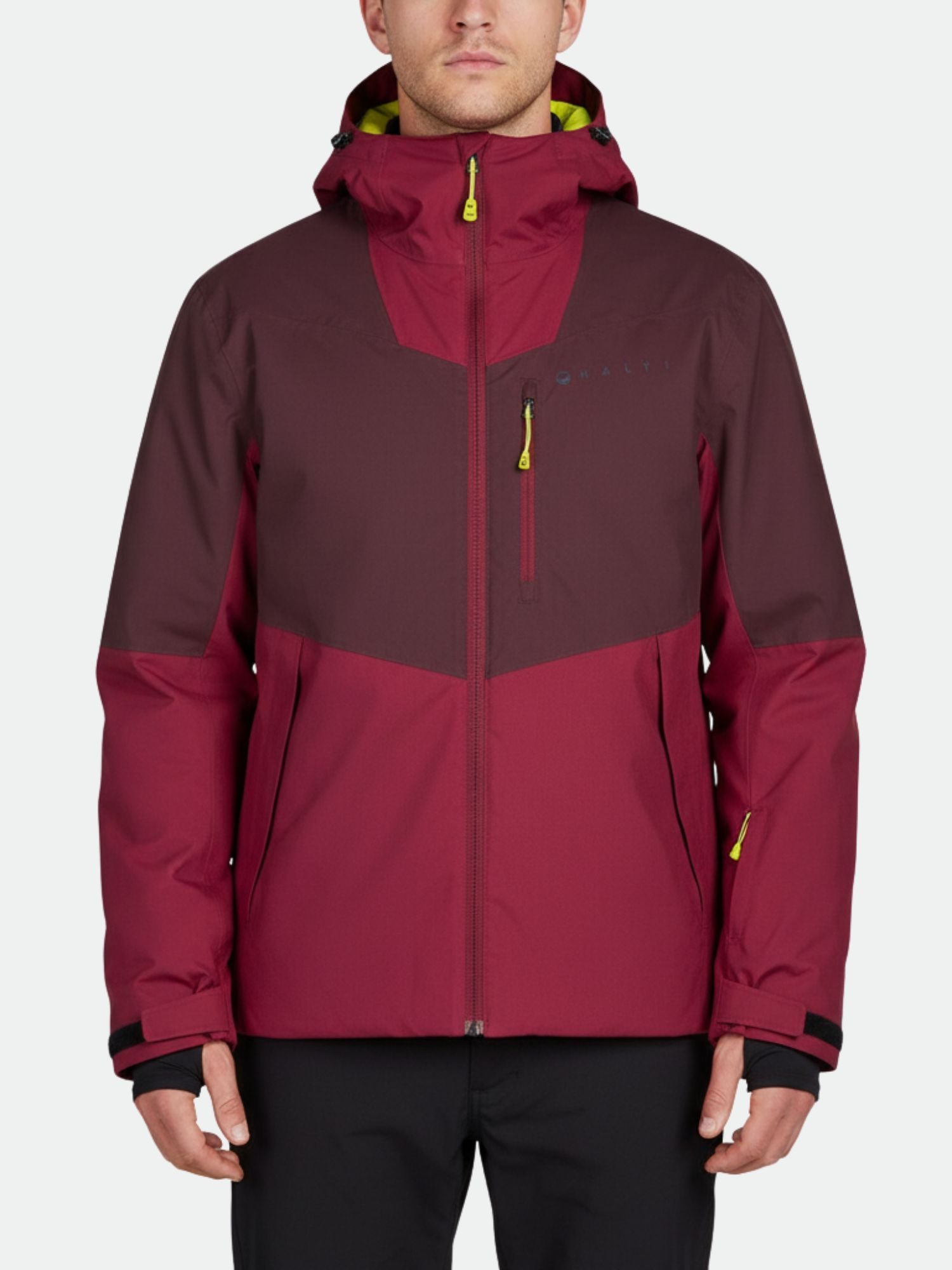 Eine Person trägt eine kastanienbraune Halti Wedeln DrymaxX Skijacke Herren mit gelben Reißverschlüssen vor einem einfarbigen Hintergrund.