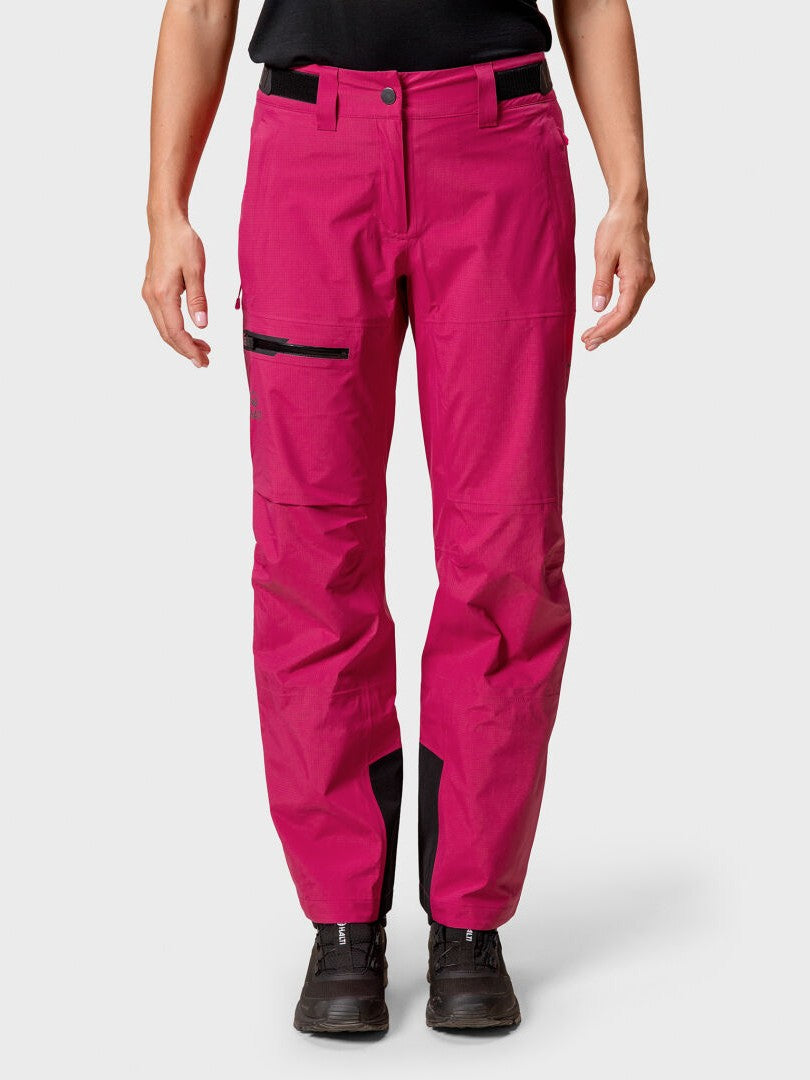 Eine Person in leuchtend rosa Halti Alpine 3L DrymaxX Skihose Damen, schwarzen Schuhen und einem schwarzen Hemd steht vor einem schlichten Hintergrund.