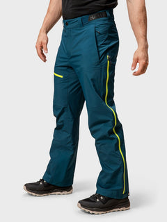 Eine Person in Halti Alpine 3L DX Skihose Herren teal Hosen mit gelben Reißverschlüssen und schwarze Wanderschuhe, auf einem unifarbenen Hintergrund.