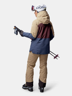 Eine Person in einer Halti Gracer DX Skijacke Damen und blauer Ausrüstung, die Skistöcke in der Hand hält, blickt vor einem weißen Hintergrund weg.