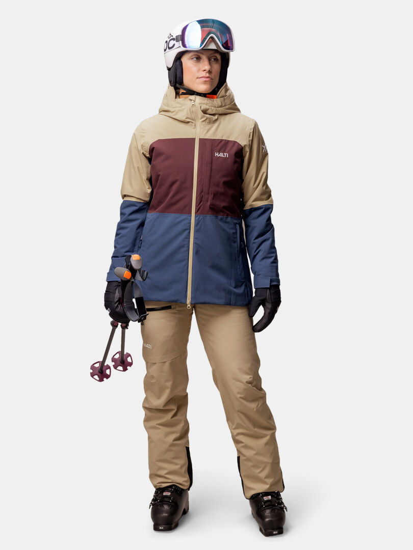 Eine Person trägt die Halti Gracer DX Skijacke Damen in beige-braun-blau, mit Helm, Skibrille und schwarzen Handschuhen.