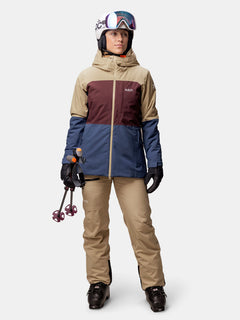 Eine Person trägt die Halti Gracer DX Skijacke Damen in beige-braun-blau, mit Helm, Skibrille und schwarzen Handschuhen.