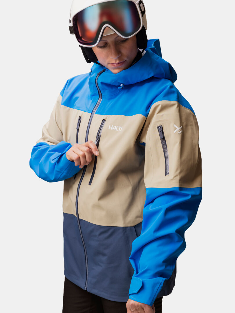 Person in einer Halti Rizle Unisex 3L DX Skijacke, die Brusttasche wird mit einem Reißverschluss geschlossen, während sie einen Skihelm trägt.