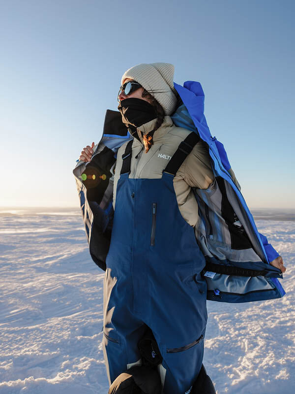 Eine Person, die eine blaue Halti Rizle Unisex 3L DX Skijacke und dazu passende Latzhosen, eine beige Mütze und eine Sonnenbrille trägt, auf einem verschneiten Berg.