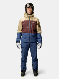 Eine Person steht vorne und trägt die Halti Melvik DX Skijacke Herren in Beige, Braun und Blau sowie eine komplette Skiausrüstung.