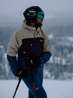 Ein Skifahrer in der Halti Melvik DX Skijacke Herren steht draußen mit Skistöcken vor einer verschneiten Landschaft.