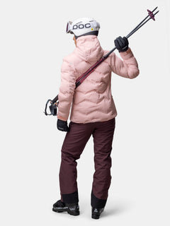 Person in Skikleidung schaut weg, hält Skier, in einer Halti Nordic Arcty II Skijacke Damen, rosa Jacke, kastanienbraune Hose, Helm.