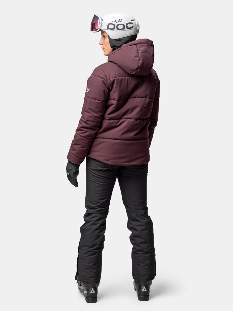 Eine Person in einer kastanienbraunen Halti Ascale Puffer Skijacke Damen und Helm steht bereit zum Skifahren auf einem weißen Hintergrund.