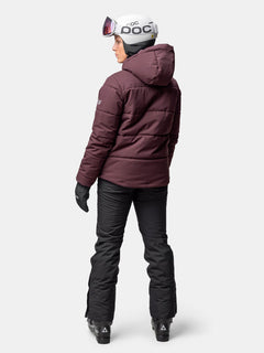 Eine Person in einer kastanienbraunen Halti Ascale Puffer Skijacke Damen und Helm steht bereit zum Skifahren auf einem weißen Hintergrund.