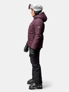 Die Frau im Profil trägt eine kastanienbraune Halti Ascale Puffer Skijacke Damen, eine schwarze Hose, Skistiefel, einen Helm und eine Schutzbrille.