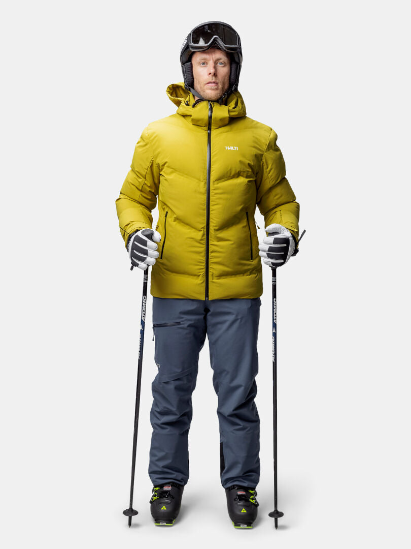 Person in der Halti Nordic Arcty II Skijacke Herren, blauen Hosen, Helm und Skistöcken auf weißem Hintergrund.