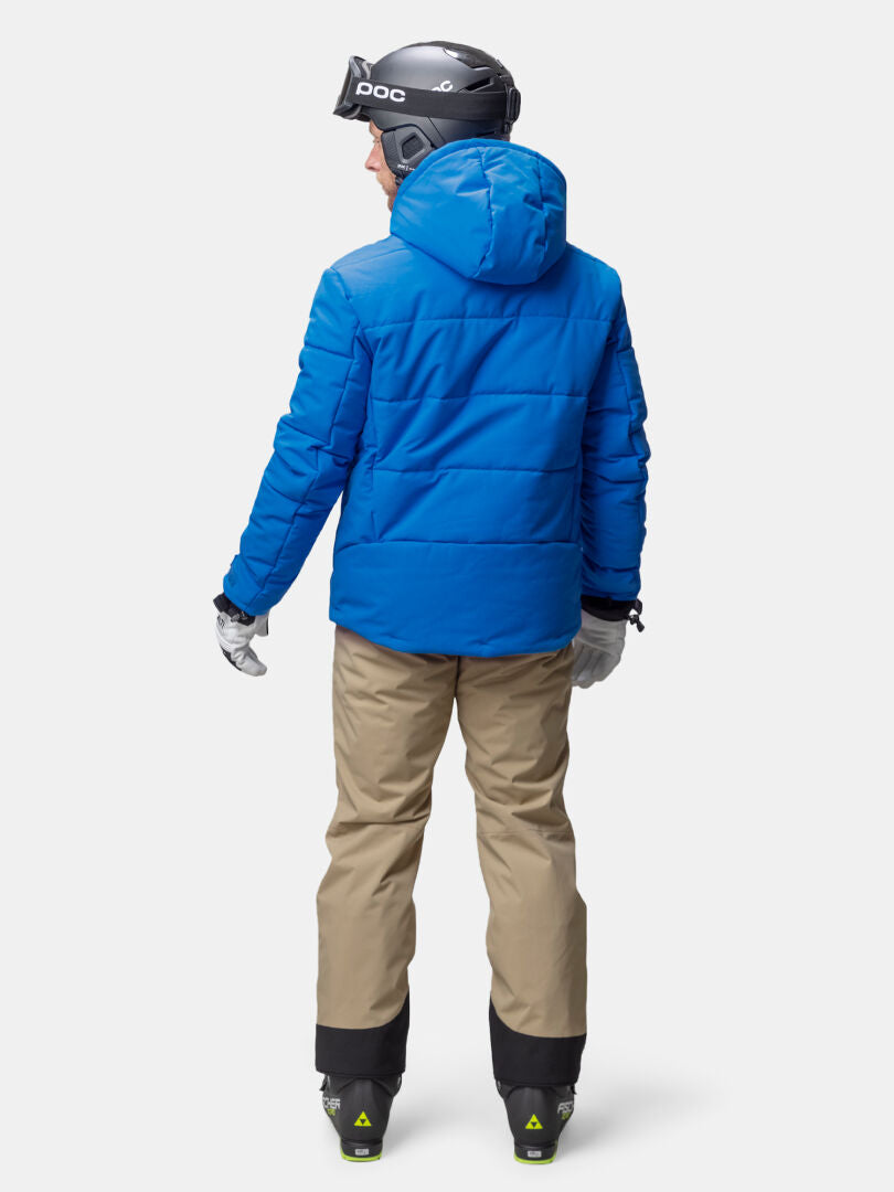 Eine Person in einer Halti Ascale Puffer Skijacke Herren, hellbrauner Hose, Handschuhen und Helm steht mit dem Gesicht zur Wand vor einem schlichten Hintergrund.