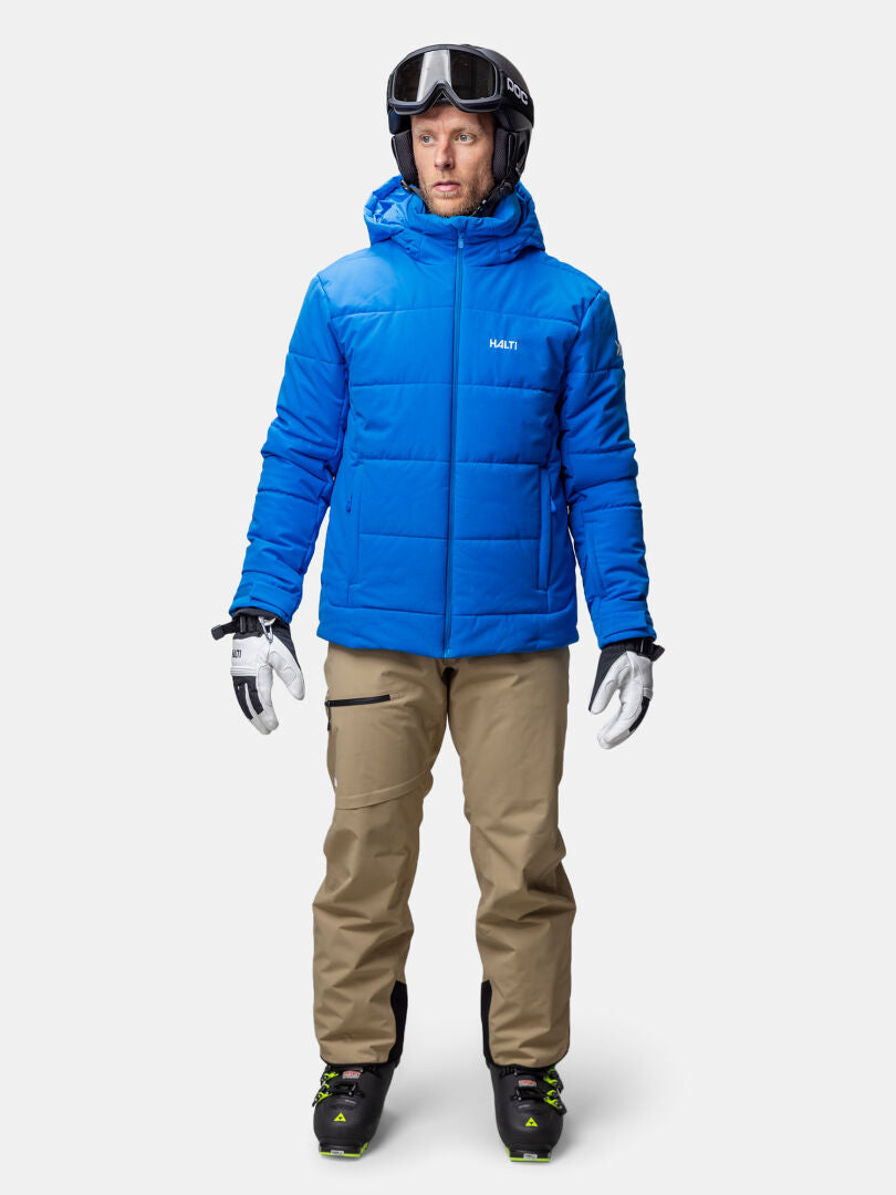 Ein Mann in einer blauen Halti Ascale Puffer Skijacke Herren, hellbrauner Hose, Handschuhen, Skistiefeln, Helm und Brille blickt nach vorne.