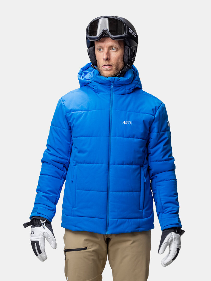 Ein Mann in einer blauen Halti Ascale Puffer Skijacke Herren, Skibrille, Handschuhen und hellbrauner Hose steht vor einem weißen Hintergrund.
