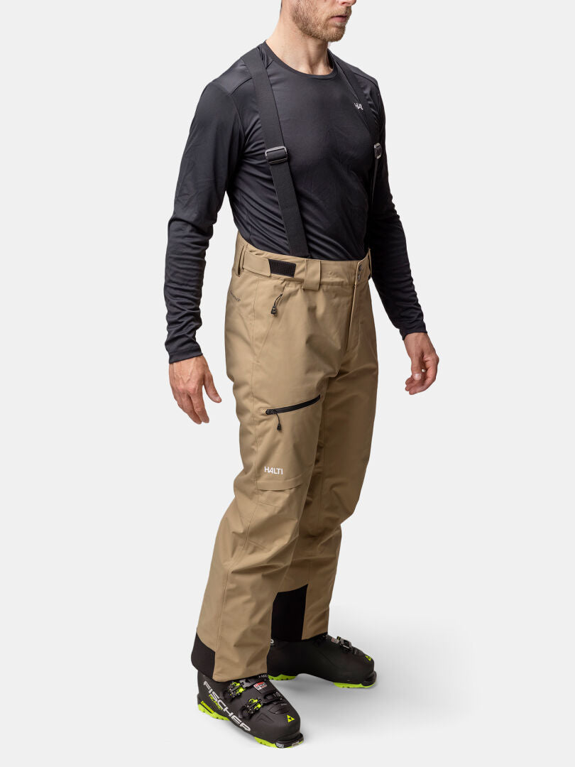 Ein Mann trägt hellbraune Halti Carvey II DX Skihose Herren mit schwarzen Hosenträgern und schwarz-grünen Wanderschuhen für den Wintersport.