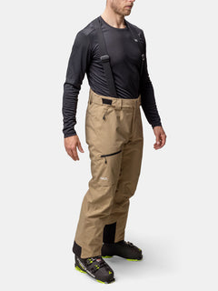 Ein Mann trägt hellbraune Halti Carvey II DX Skihose Herren mit schwarzen Hosenträgern und schwarz-grünen Wanderschuhen für den Wintersport.
