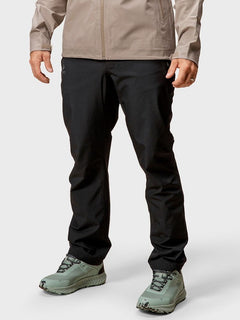 Eine Person in beiger Jacke, Halti Vuoksi Herren Recy DrymaxX Long Shellhose und hellgrünen Trailschuhen steht vor einem schlichten Hintergrund.