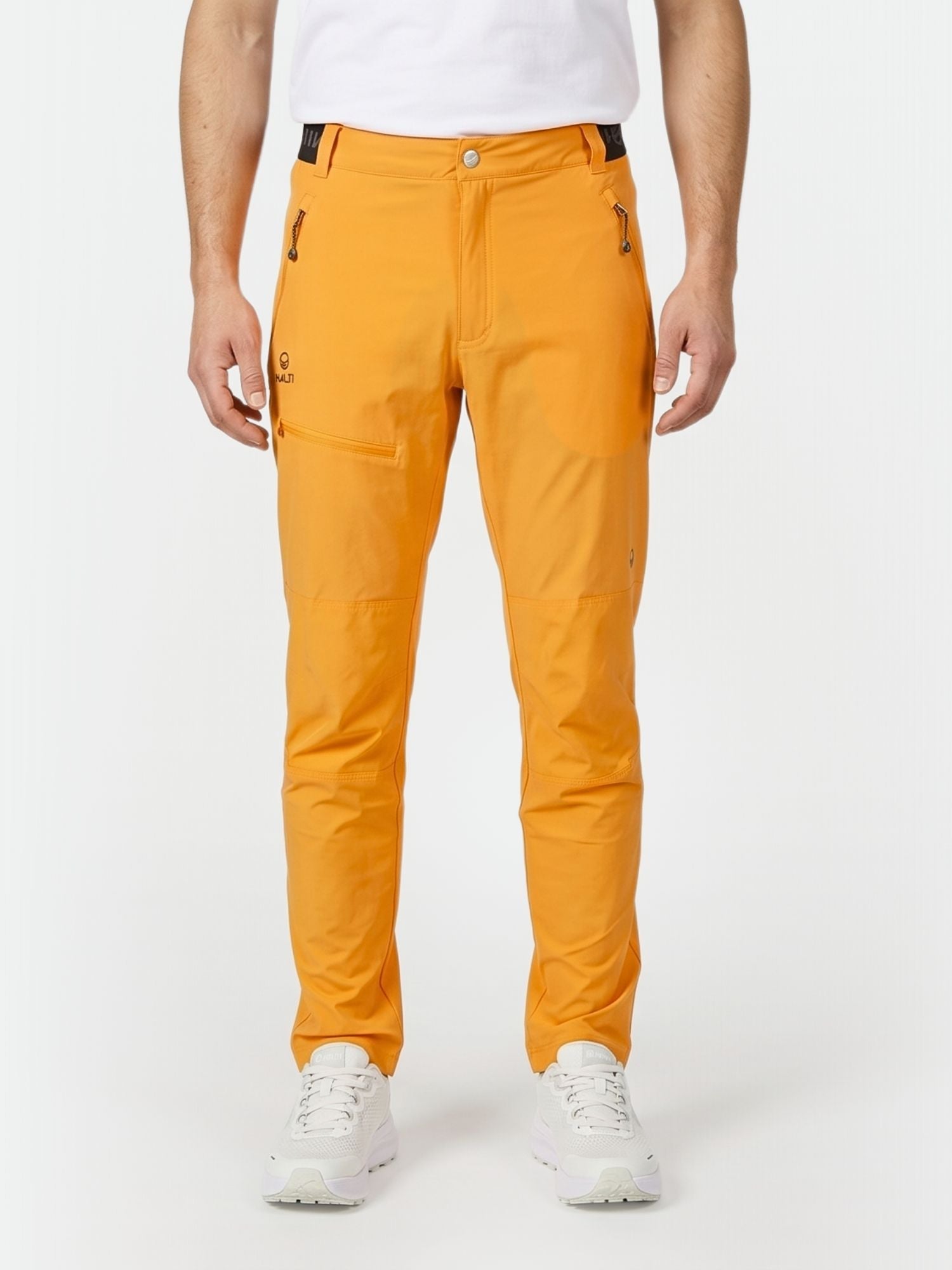 Eine Person in leuchtend orangefarbener Halti Pallas Herren X-stretch Lite Hose, einem weißen Hemd und Turnschuhen steht vor einem weißen Hintergrund.
