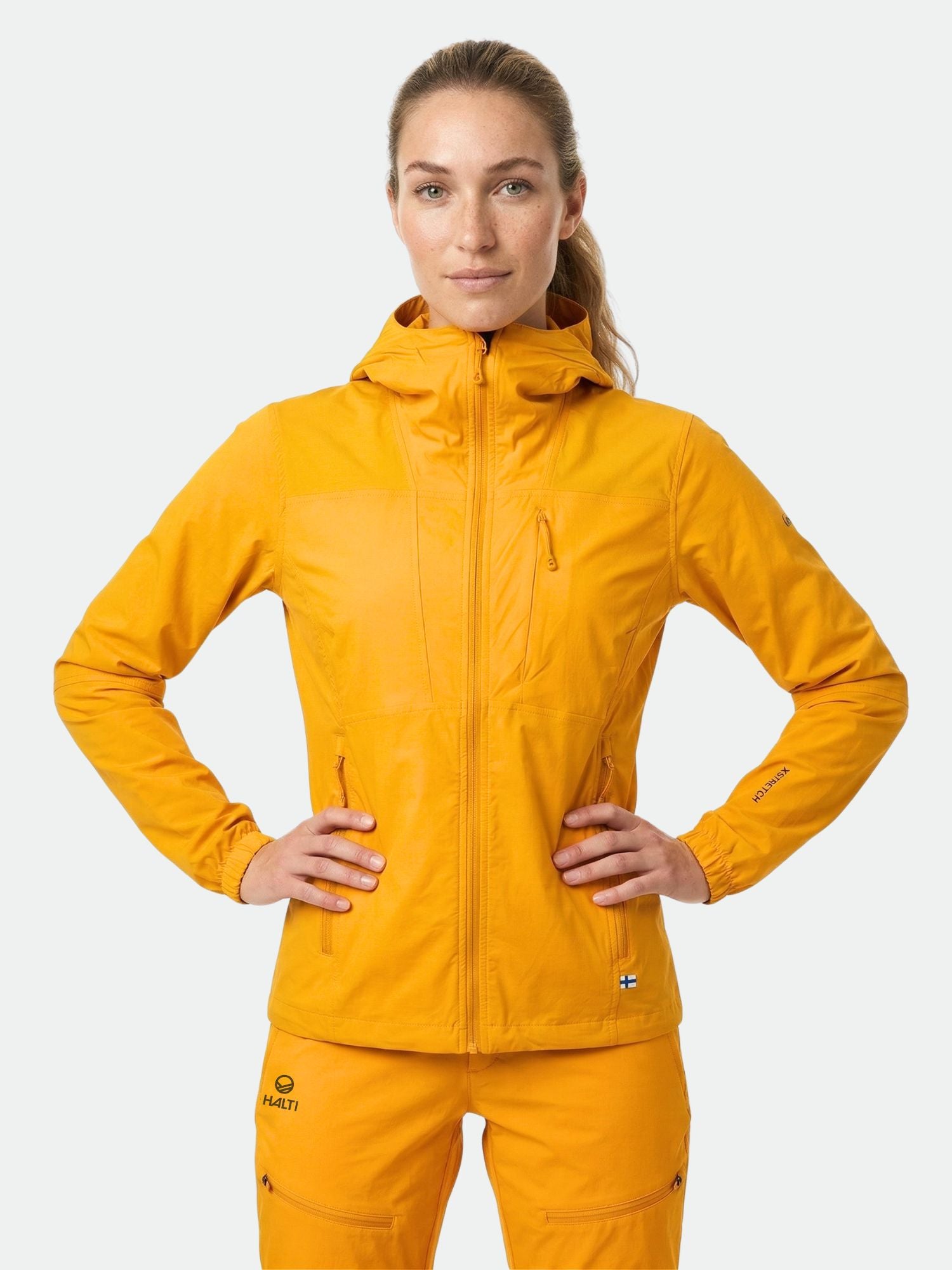 Eine Frau in einer leuchtend orangefarbenen Halti Pallas II Damen X-stretch Jacke und passender Hose, die Hände in die Hüften gestemmt, schaut nach vorne.