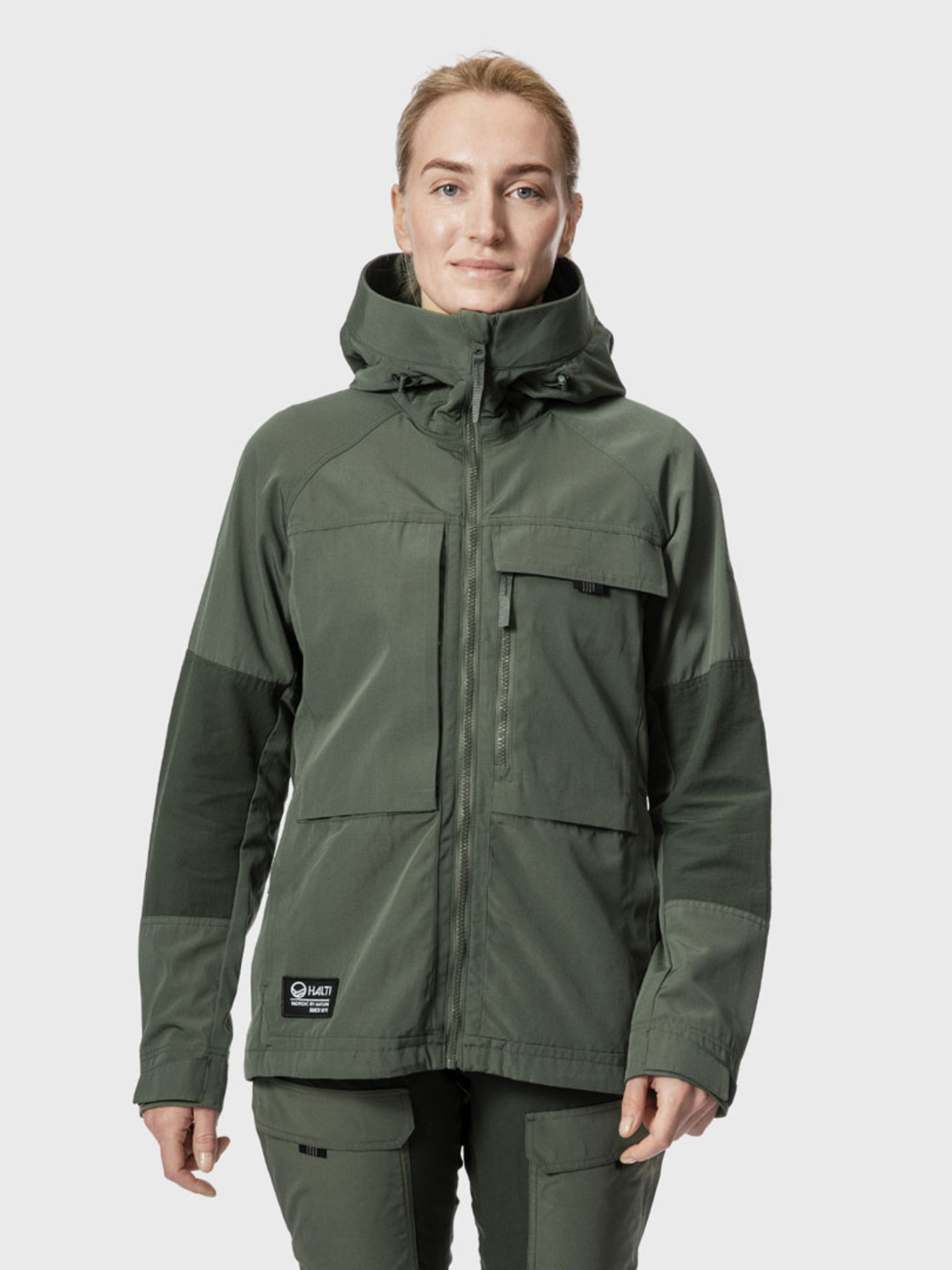 Eine Person trägt die Halti Hiker Damen Ventilated Jacke und die dazu passende Hose und schaut in die Kamera vor einem schlichten Hintergrund.