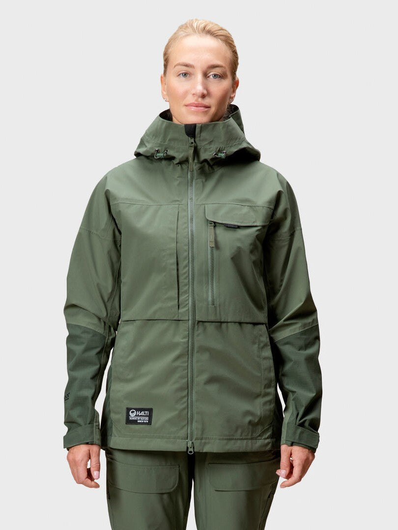 Eine Frau in einer grünen Halti Hiker DrymaxX 3L Ventilated Jacke steht vor einem schlichten hellgrauen Hintergrund.