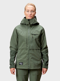 Eine Frau in einer grünen Halti Hiker DrymaxX 3L Ventilated Jacke steht vor einem schlichten hellgrauen Hintergrund.