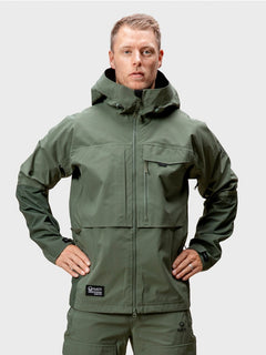 Ein Mann schaut nach vorne, er trägt eine grüne Halti Hiker DrymaxX 3L Ventilated Jacke Herren und eine passende Hose, die Hände in die Hüften gestemmt.