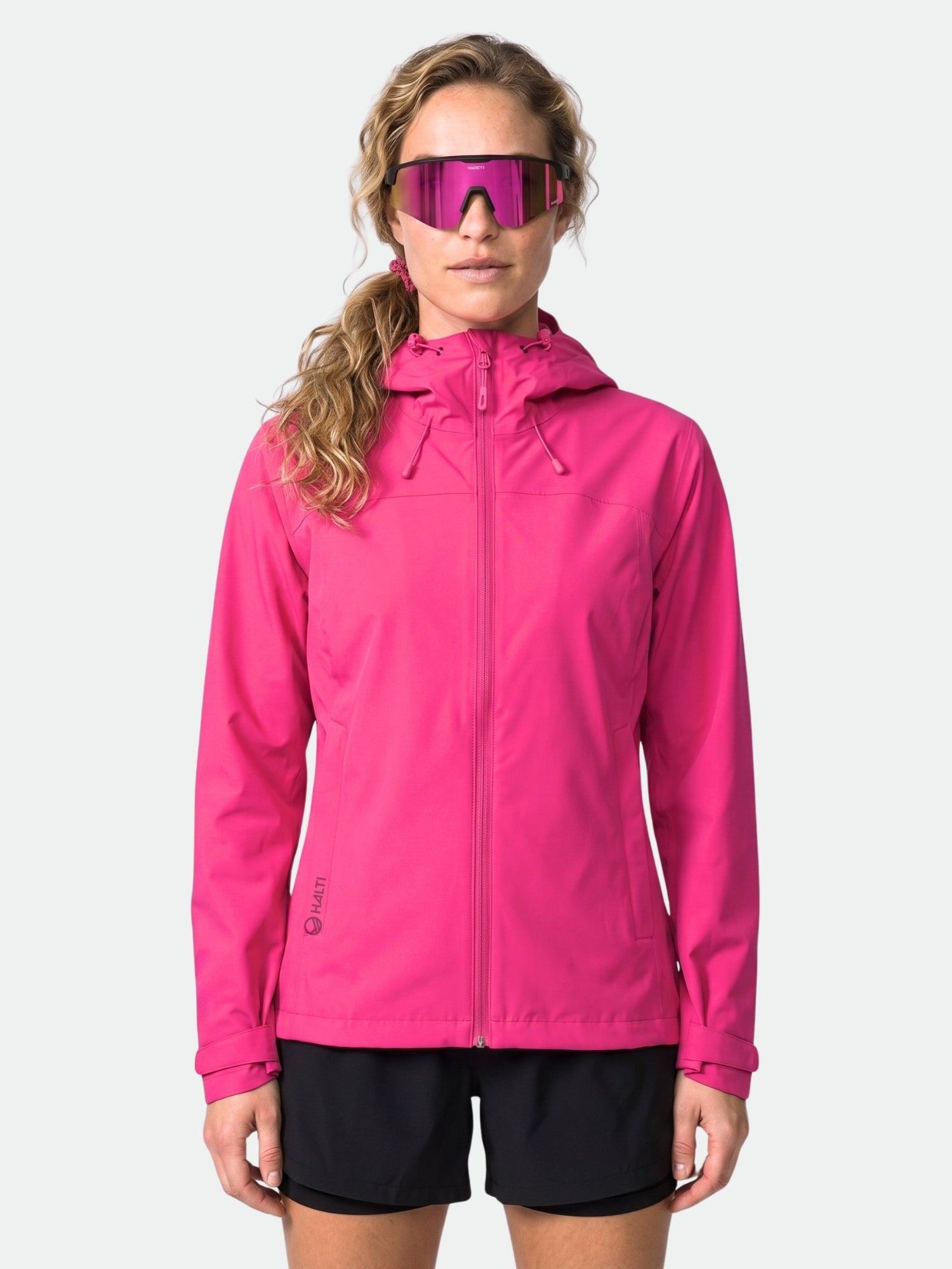 Eine Person trägt die Halti Alma Damen DX Regenjacke in leuchtendem Pink und schwarze Shorts und steht mit dem Gesicht nach vorne.