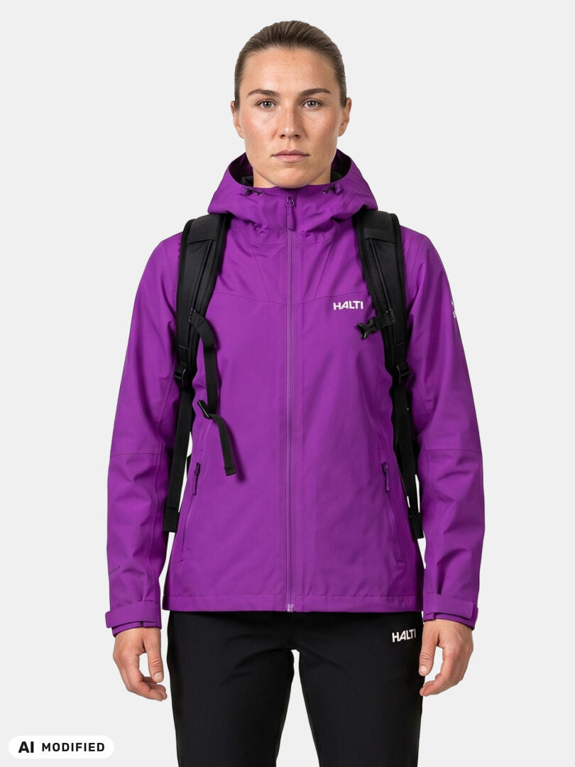 Eine Frau in einer „Halti Forter DX Regenjacke“ für Damen, mit schwarzem Rucksack und Hose, bereit zum Wandern vor einem einfarbigen Hintergrund.