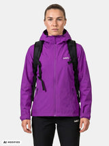 Eine Frau in einer „Halti Forter DX Regenjacke“ für Damen, mit schwarzem Rucksack und Hose, bereit zum Wandern vor einem einfarbigen Hintergrund.