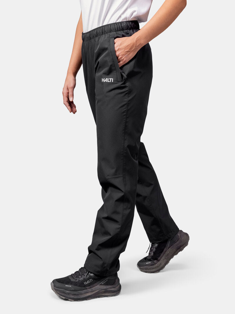 Person, die schwarze Halti Forter DX Regenhose Damen, schwarze Turnschuhe und ein weißes Hemd trägt und mit einer Hand in der Tasche geht.