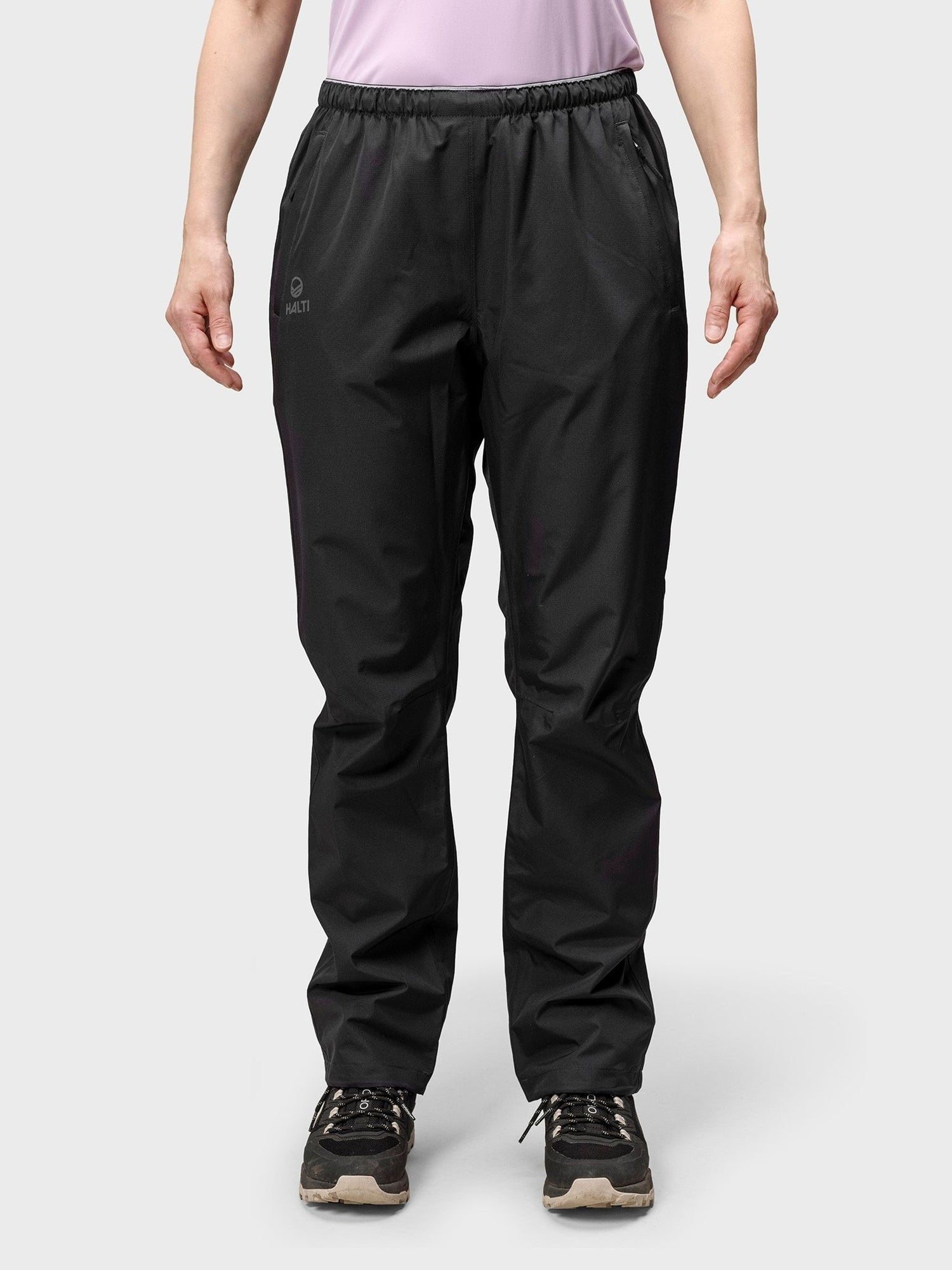 Eine Person trägt Halti Forter DX Regenhose Damen in Schwarz, schwarze Turnschuhe und ein hellviolettes Hemd vor einem schlichten Hintergrund.