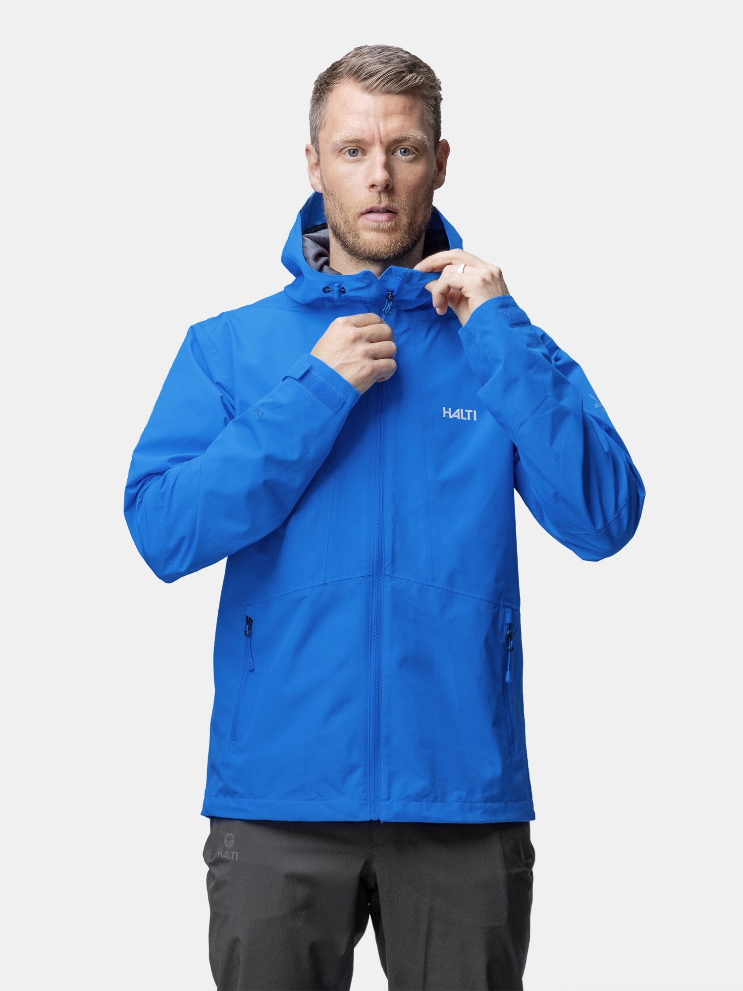 Ein Mann in einer leuchtend blauen Halti Forter DX Regenjacke Herren zieht den Reißverschluss hoch, während er vor einem schlichten Hintergrund nach vorne schaut.