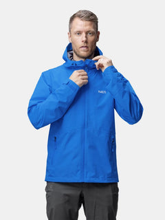 Ein Mann in einer blauen Halti Forter DX Regenjacke Herren schließt den Reißverschluss seiner wetterfesten Outdoor-Jacke und schaut dabei nach vorne.