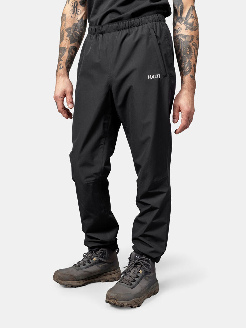 Eine Person in schwarzer Halti Forter DrymaxX Regenhose Herren und Wanderschuhen steht vor einem schlichten weißen Hintergrund.