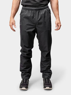 Eine Person in Halti Forter DX Regenhose Herren und Turnschuhen steht vor einem hellen Hintergrund, bereit zum Wandern oder Regen.