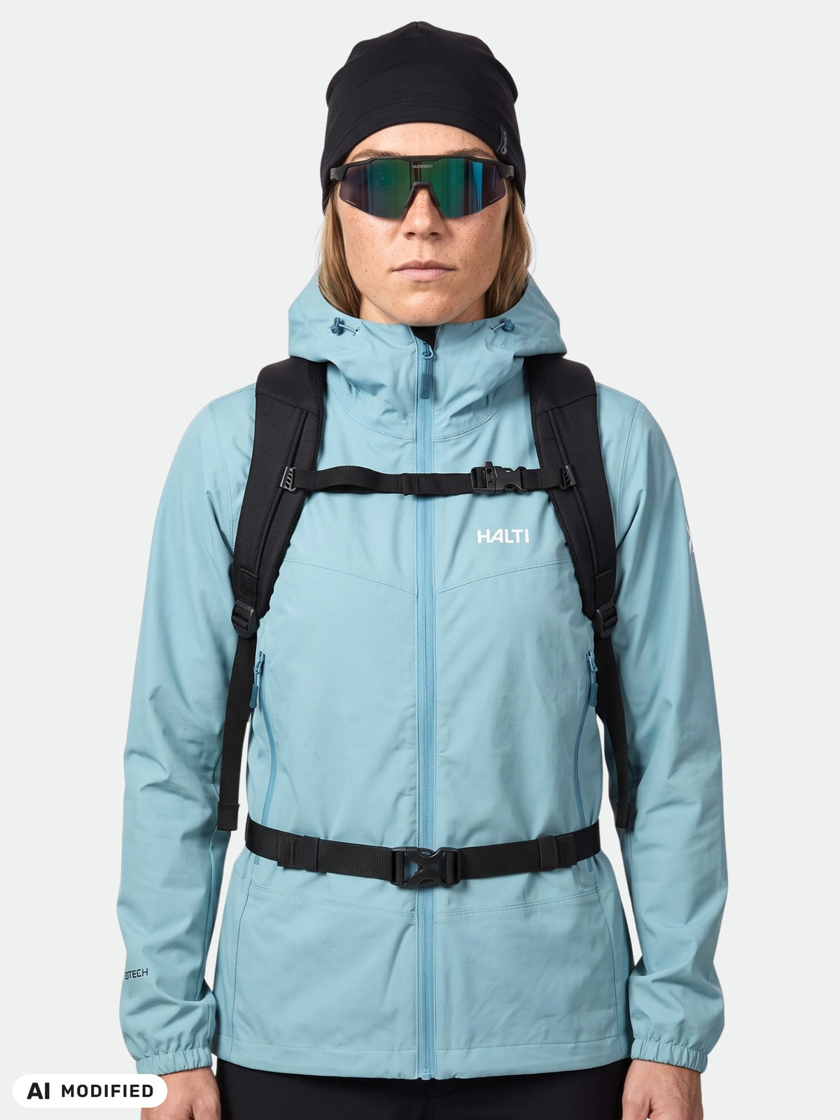 Person, die eine blaue Halti Pallas Evo X-Stretch-Jacke für Damen, eine schwarze Mütze, eine Sonnenbrille und einen Rucksack trägt, vor weißem Hintergrund.