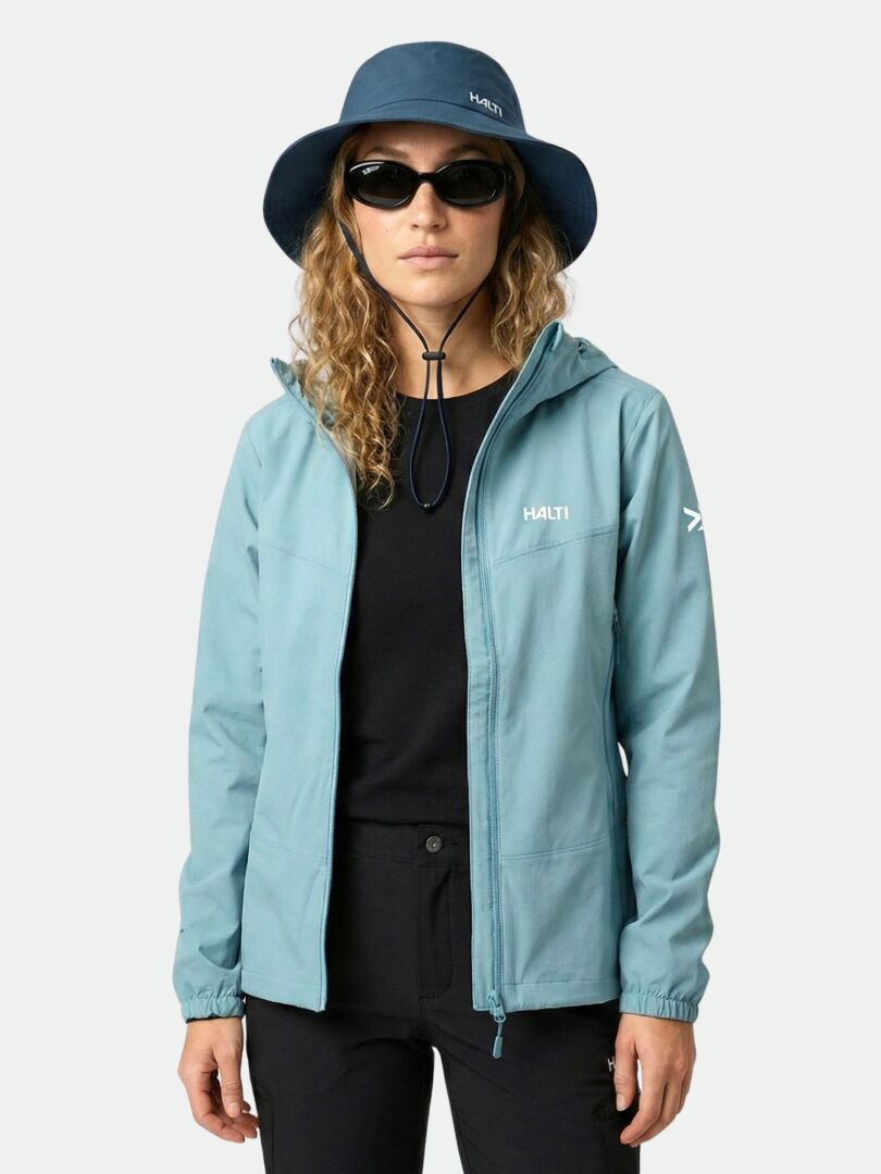 Eine Frau in einer Halti Pallas Evo X-Stretch-Jacke Damen, blauem Hut und Sonnenbrille steht mit dem Gesicht nach vorne vor einem schlichten Hintergrund.