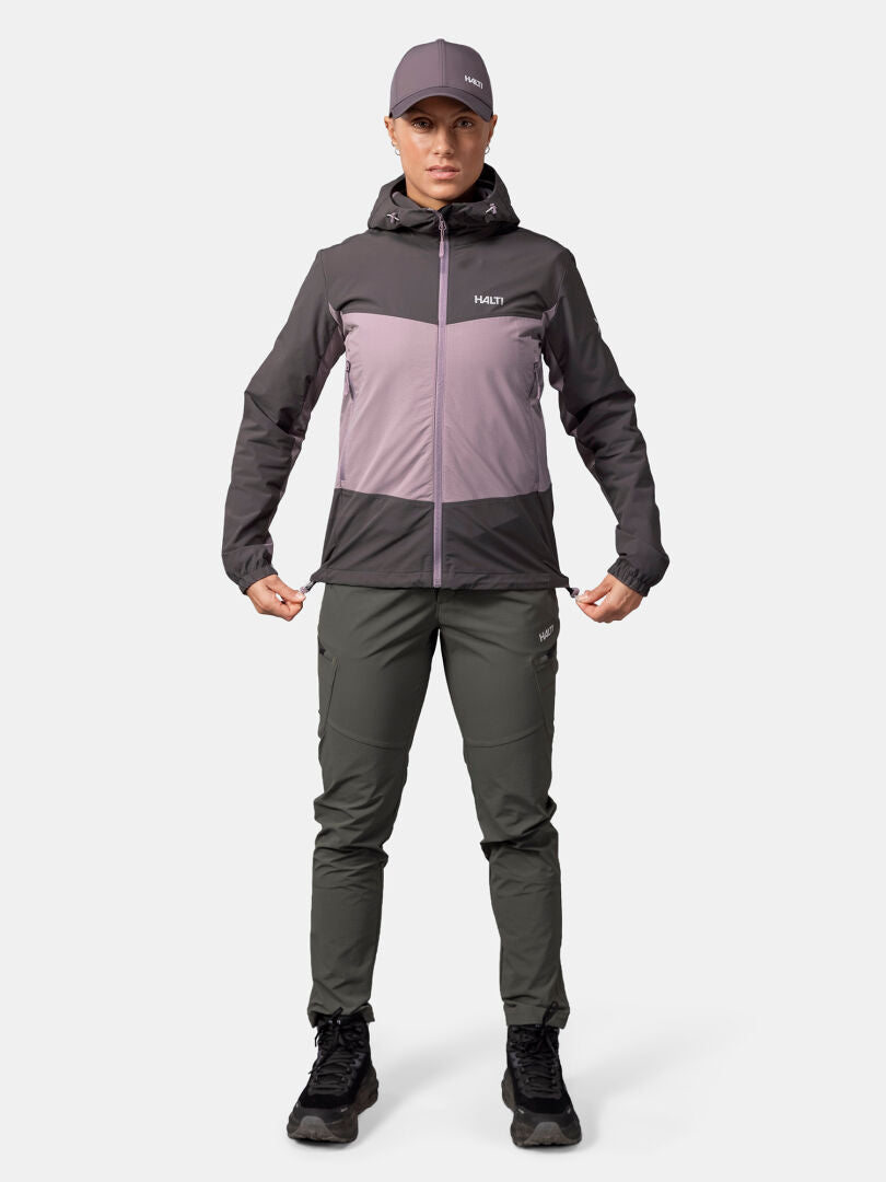 Eine Person trägt eine lilafarbene Halti Pallas Evo X-Stretch-Jacke Damen, eine graue Hose und eine lilafarbene Mütze und schaut nach vorne.