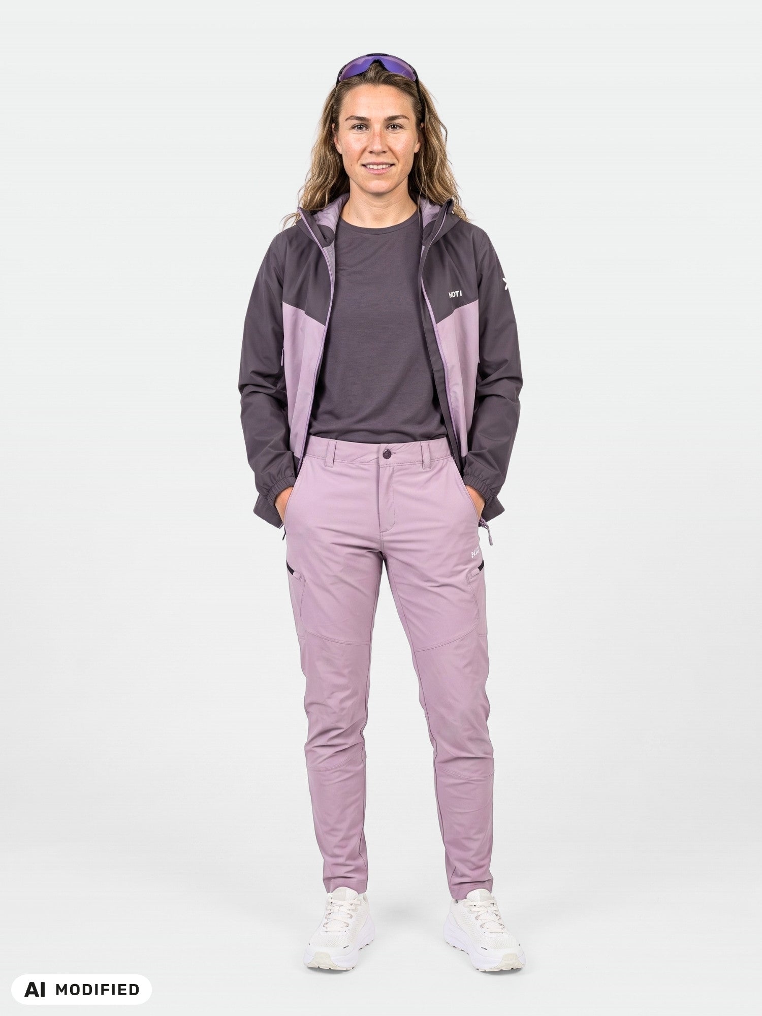 Stehende Person, bekleidet mit einer grauen Jacke, einer „Halti Pallas Evo X-Stretch“-Hose für Damen in Lila, weißen Schuhen und einer Sonnenbrille auf dem Kopf.