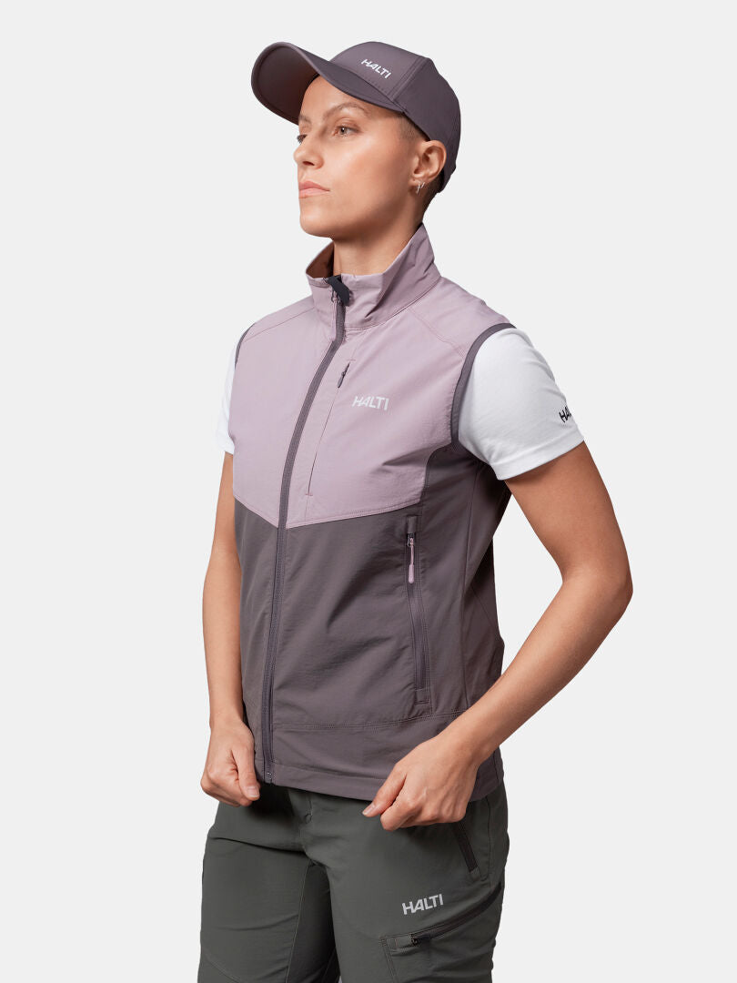 Die Person trägt eine lila Halti-Mütze, eine Halti Pallas Evo X-stretch Weste Damen, ein weißes T-Shirt und eine grüne Halti-Hose.