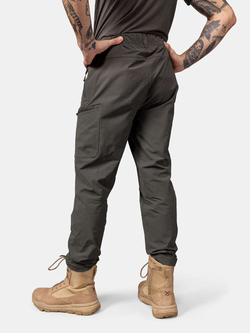 Person trägt Halti Pallas Evo X-Stretch Hosen Herren in oliv, hellbraune Stiefel, Rücken zur Kamera, Hände in die Hüften gestemmt, Armtattoos.