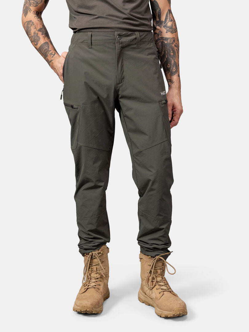 Ein Mann trägt eine Halti Pallas Evo X-Stretch Hose in Olivgrün, ein schwarzes Hemd und hellbraune Stiefel vor einem schlichten Hintergrund.