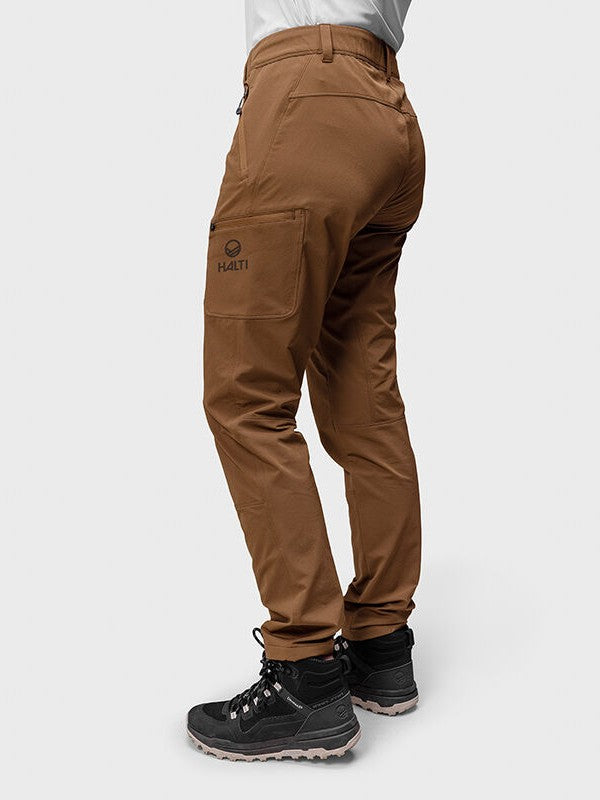 Frau in brauner Halti Pallas Evo Brushed X-Stretch-Hose und schwarzen Wanderschuhen, seitlich stehend auf weißem Hintergrund.