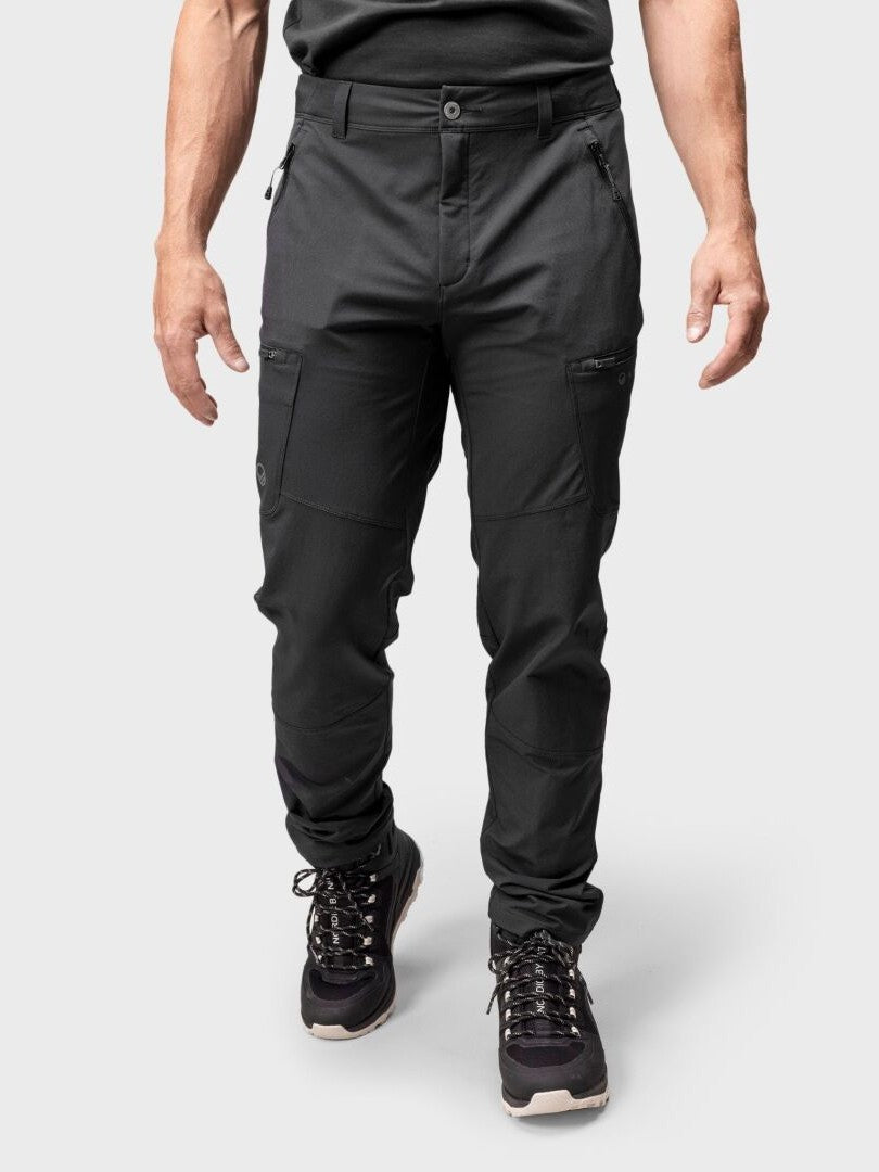 Eine Person trägt Halti Pallas Evo Brushed X-stretch Hosen Herren und schwarze Wanderschuhe vor einem schlichten hellgrauen Hintergrund.