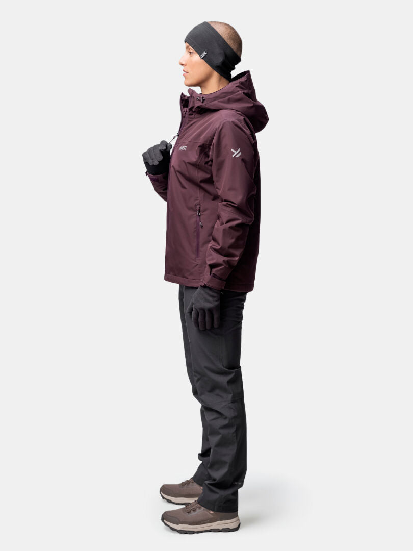 Seitlich stehende Frau in einer kastanienbraunen Halti Forter DX Warm Regenjacke Damen, schwarzer Hose und braunen Wanderstiefeln.