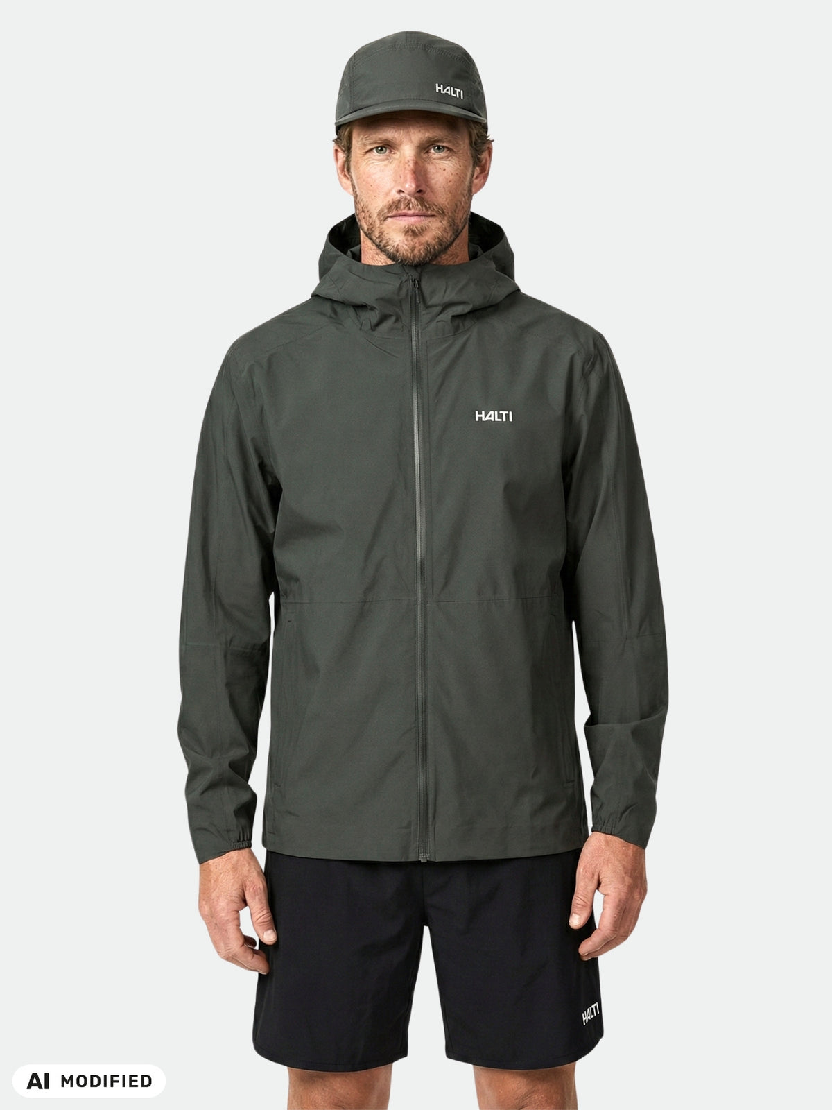 Ein Mann trägt vor einem einfarbigen Hintergrund eine Halti Pisarat 2,5L DX Regenjacke für Herren mit DrymaxX 2 Tech, schwarze Shorts und eine Mütze.